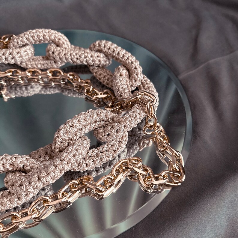 May include: A close-up of a beige crocheted chain necklace with a gold chain interwoven. The necklace is lying on a reflective surface.