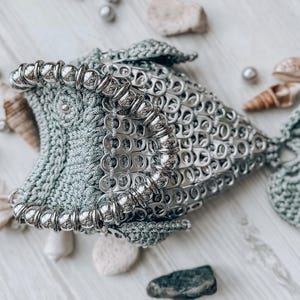 以下が含まれることがあります： 銀色の金属タブと薄緑色の糸で作られた手作りの魚の彫刻。 魚はかぎ針編みの体と尾を持つ詳細なデザインで、装飾的なビーズで飾られています。 彫刻は貝殻と真珠と一緒に展示されています。
