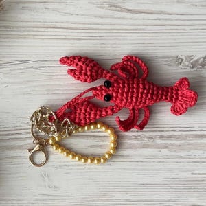 Puede incluir: Un llavero de langosta de crochet rojo con ojos negros y una cadena dorada. La langosta est&aacute; unida a una cadena dorada con un brazalete de perlas.