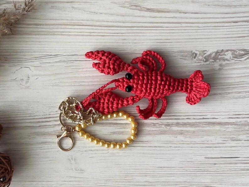 Puede incluir: Un llavero de langosta de crochet rojo con ojos negros y una cadena dorada. La langosta est&aacute; unida a una cadena dorada con un brazalete de perlas.