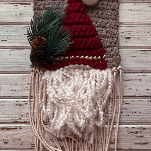 May include: A woven wall hanging featuring a red Santa hat with a white pom-pom, a green pine branch with a pine cone, and a white yarn beard. The background is a gray woven fabric.