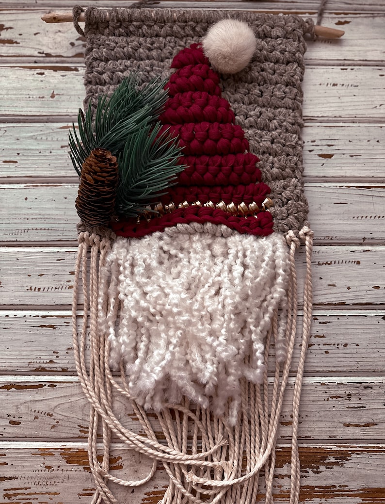 May include: A woven wall hanging featuring a red Santa hat with a white pom-pom, a green pine branch with a pine cone, and a white yarn beard. The background is a gray woven fabric.