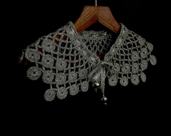 May include: A silver crochet collar with a delicate, lacy design. The collar has a tie at the front and is hanging on a wooden hanger.