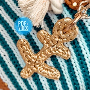 May include: A gold-colored crocheted anchor charm with a white and teal striped background. The anchor is attached to a silver ring.