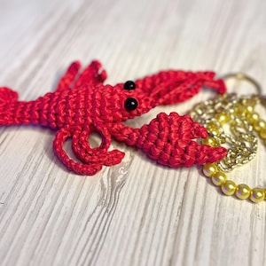 Puede incluir: Un llavero de langosta rojo tejido a crochet con ojos negros y una cadena dorada con cuentas amarillas.