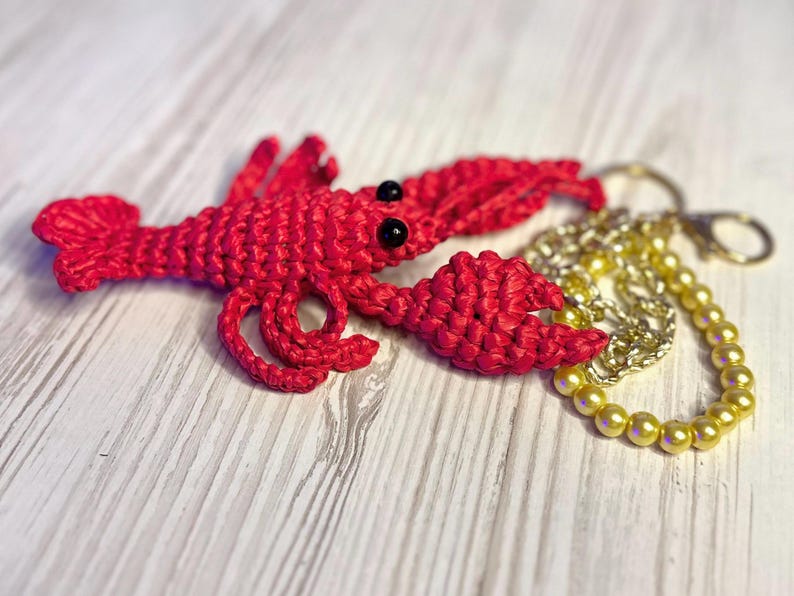Puede incluir: Un llavero de langosta rojo tejido a crochet con ojos negros y una cadena dorada con cuentas amarillas.