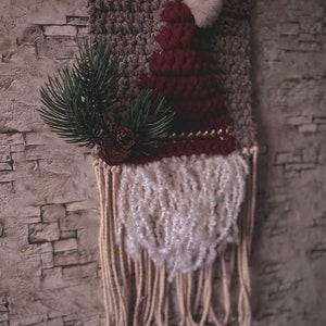 May include: A gray and red knitted wall hanging with a white pom-pom on top. The hanging has a white fringe at the bottom and a green pine branch with a pine cone.