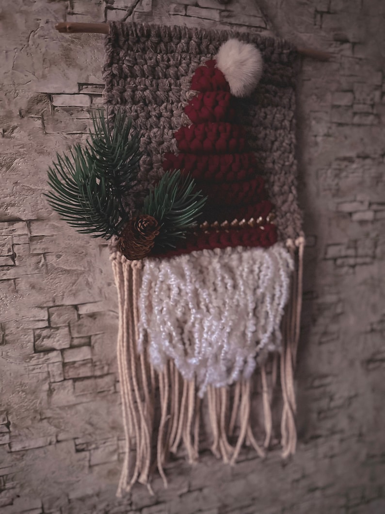 May include: A gray and red knitted wall hanging with a white pom-pom on top. The hanging has a white fringe at the bottom and a green pine branch with a pine cone.