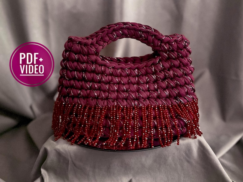 May include: A burgundy crochet handbag with a handle and beaded fringe. The bag is made of thick, textured yarn with silver threads woven throughout. The fringe is made of small, red beads.
