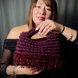 May include: A handmade, dark purple handbag with a textured, woven design. The bag features a solid handle and a fringe of dark red beads along the bottom edge. The bag is held by a person wearing a black dress.