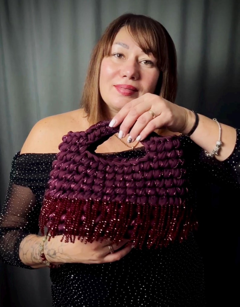 May include: A handmade, dark purple handbag with a textured, woven design. The bag features a solid handle and a fringe of dark red beads along the bottom edge. The bag is held by a person wearing a black dress.