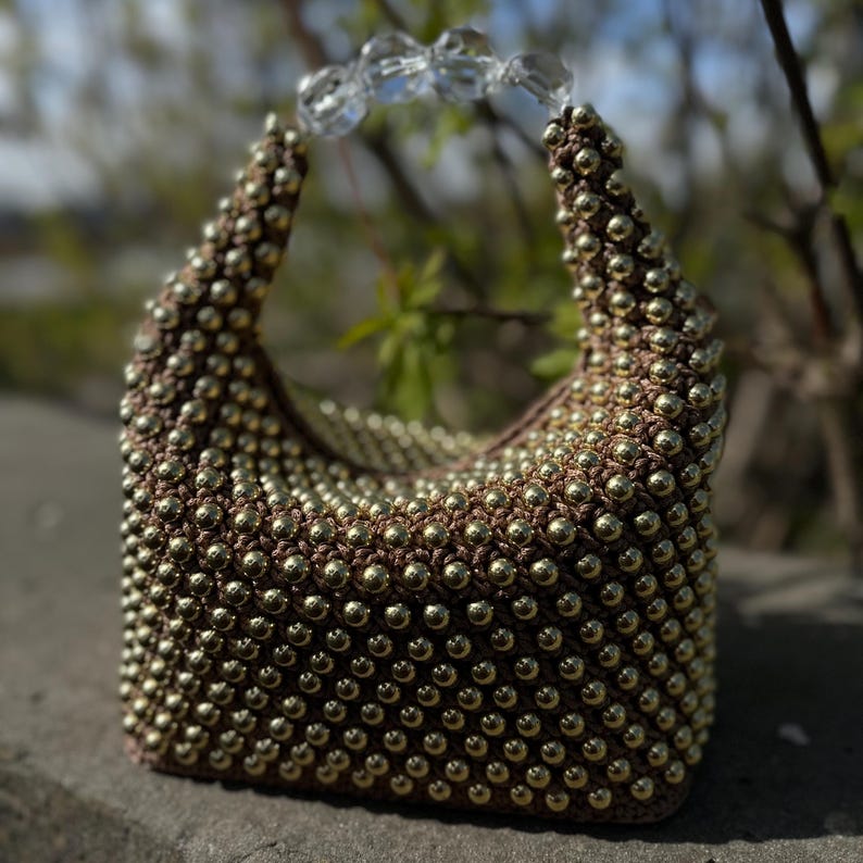 May include: A small, brown beaded handbag with a clear plastic handle. The bag is made of a woven fabric with gold beads.