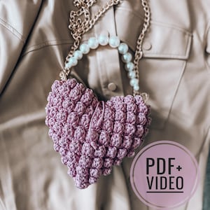 Puede incluir: Un bolso de mano en forma de corazón, tejido a ganchillo en color lavanda. Presenta un diseño texturizado de punto de burbuja y una correa de cadena con detalles de perlas. La imagen incluye un gráfico circular con el texto "PDF+VIDEO".