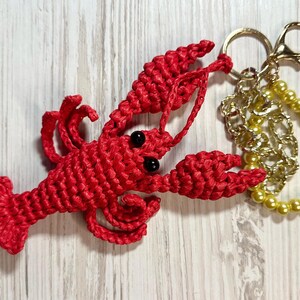 Puede incluir: Un llavero de langosta rojo tejido a crochet con ojos negros, unido a una cadena dorada y un c&iacute;rculo de cuentas doradas.