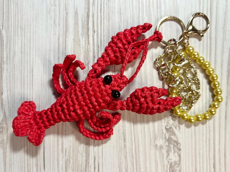 Puede incluir: Un llavero de langosta rojo tejido a crochet con ojos negros, unido a una cadena dorada y un c&iacute;rculo de cuentas doradas.
