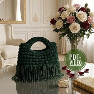 May include: A dark green, hand-crocheted handbag with a rounded handle and beaded fringe. The bag sits on a reflective surface, next to a vase of roses and a decorative book. The image includes a "PDF + VIDEO" graphic.