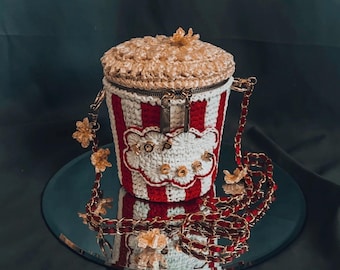 May include: A unique popcorn-shaped handbag with a crocheted design. The bag features red and white stripes, a popcorn-like top, and the word "POP CORN" embroidered on the front. It has a gold chain strap with decorative floral accents.