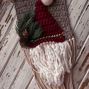 May include: A woven wall hanging featuring a Santa Claus design. The hanging is made with brown, red, and white yarn and has a white pom-pom on top. The beard is made of long white yarn strands.