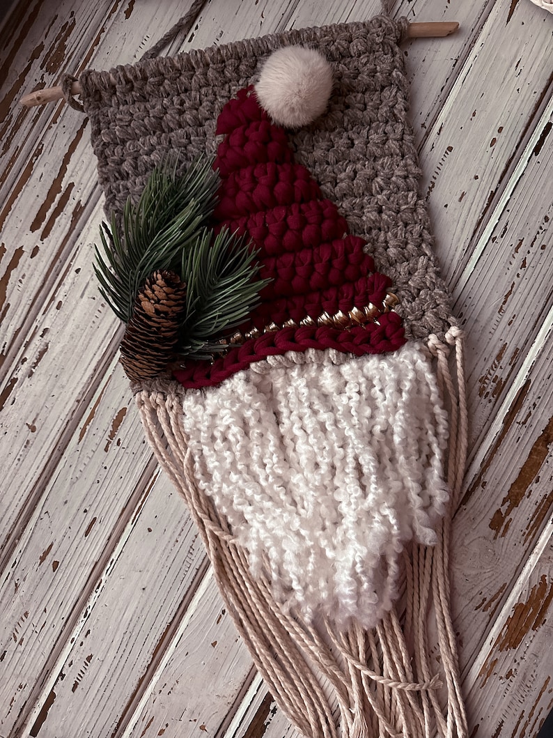 May include: A woven wall hanging featuring a Santa Claus design. The hanging is made with brown, red, and white yarn and has a white pom-pom on top. The beard is made of long white yarn strands.