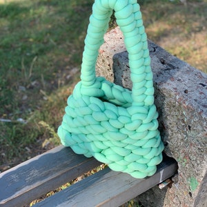 May include: A light green knitted tote bag with a braided handle. The bag is sitting on a wooden bench.