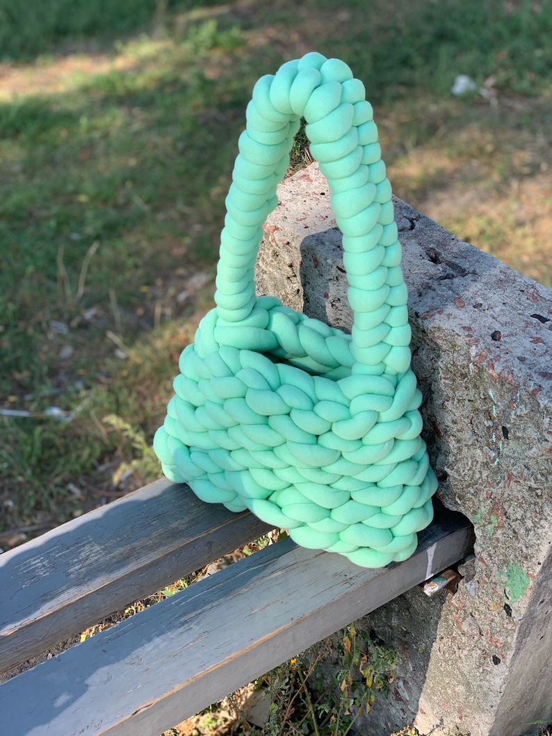 May include: A light green knitted tote bag with a braided handle. The bag is sitting on a wooden bench.