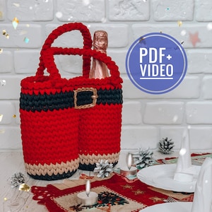 Puede incluir: Un porta botellas de crochet rojo y negro con forma de pantalón de Papá Noel con un cinturón marrón. El porta botellas contiene una botella de champán. La imagen también incluye un mantel individual rojo y blanco con un diseño de árbol de Navidad, dos platos blancos y dos velas blancas.