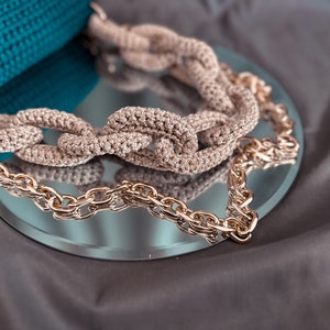 May include: A close-up of a crocheted chain bag strap with a gold chain. The strap is beige and the chain is gold. The strap is on a mirror surface.