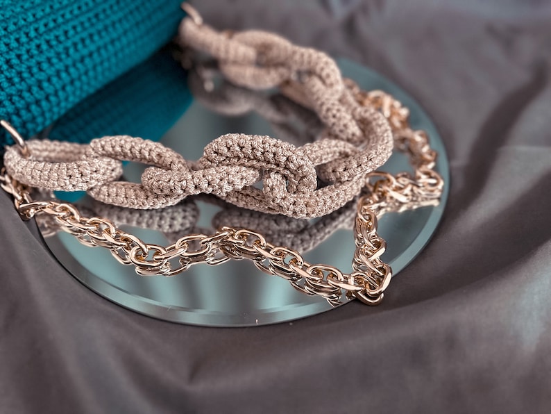 May include: A close-up of a crocheted chain bag strap with a gold chain. The strap is beige and the chain is gold. The strap is on a mirror surface.