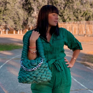 May include: A woman wearing a green jumpsuit with a matching green knitted tote bag with a silver chain handle.