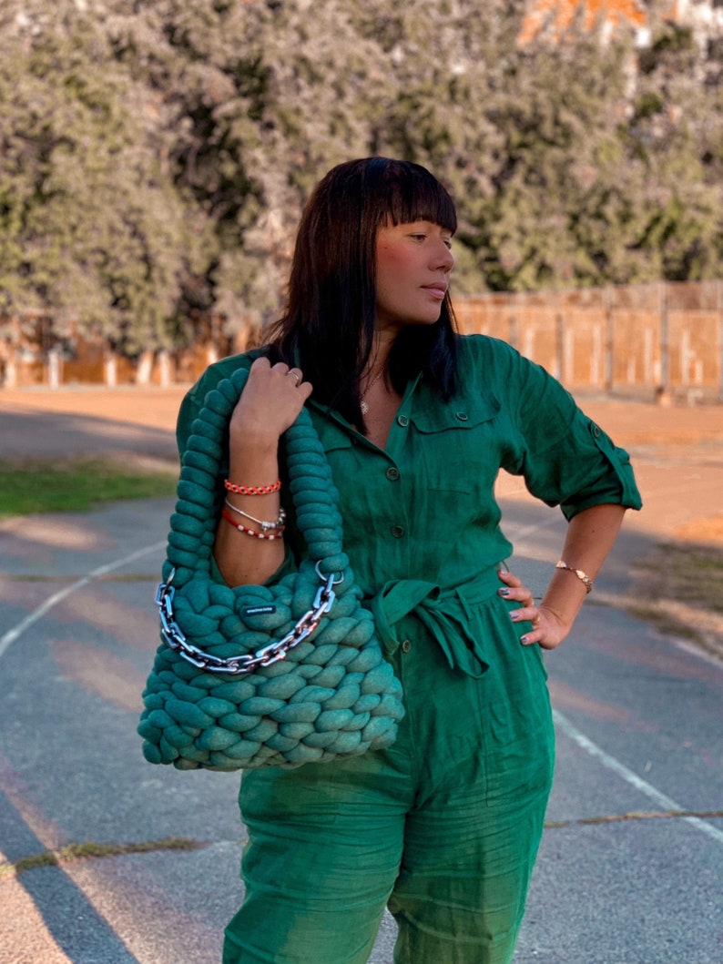 May include: A woman wearing a green jumpsuit with a matching green knitted tote bag with a silver chain handle.