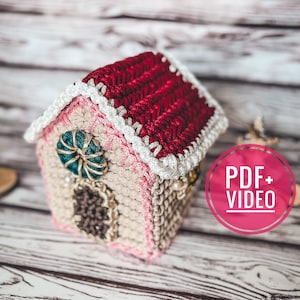May include: A handcrafted crochet house with a red roof, white trim, and a teal and gold accent. The house is beige and pink with a decorative window and door. The image includes a "PDF + VIDEO" graphic.