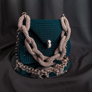 May include: A teal green crocheted handbag with a chunky chain strap. The bag has a flap closure with a decorative silver skull-shaped button.