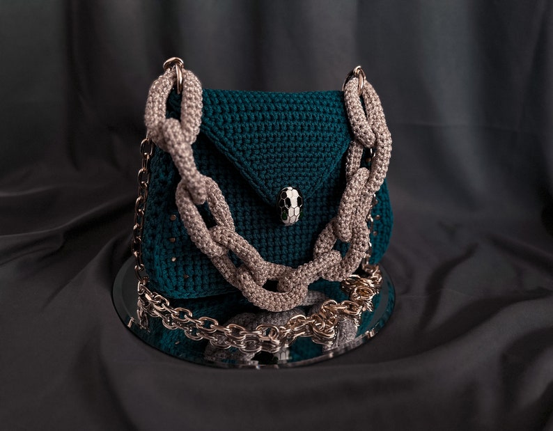 May include: A teal green crocheted handbag with a chunky chain strap. The bag has a flap closure with a decorative silver skull-shaped button.