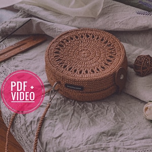 Puede incluir: Un bolso redondo de crochet marrón con una correa de cuero y un diseño circular. El bolso está sobre una superficie de tela gris claro con una superficie de madera en el fondo. El texto "PDF + VIDEO" está en un círculo rosa.