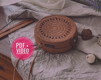 Può includere: Una borsa rotonda di colore marrone realizzata a uncinetto con una tracolla in pelle e un design circolare. La borsa è su una superficie di tessuto grigio chiaro con una superficie di legno sullo sfondo. Il testo "PDF + VIDEO" è su un cerchio rosa.
