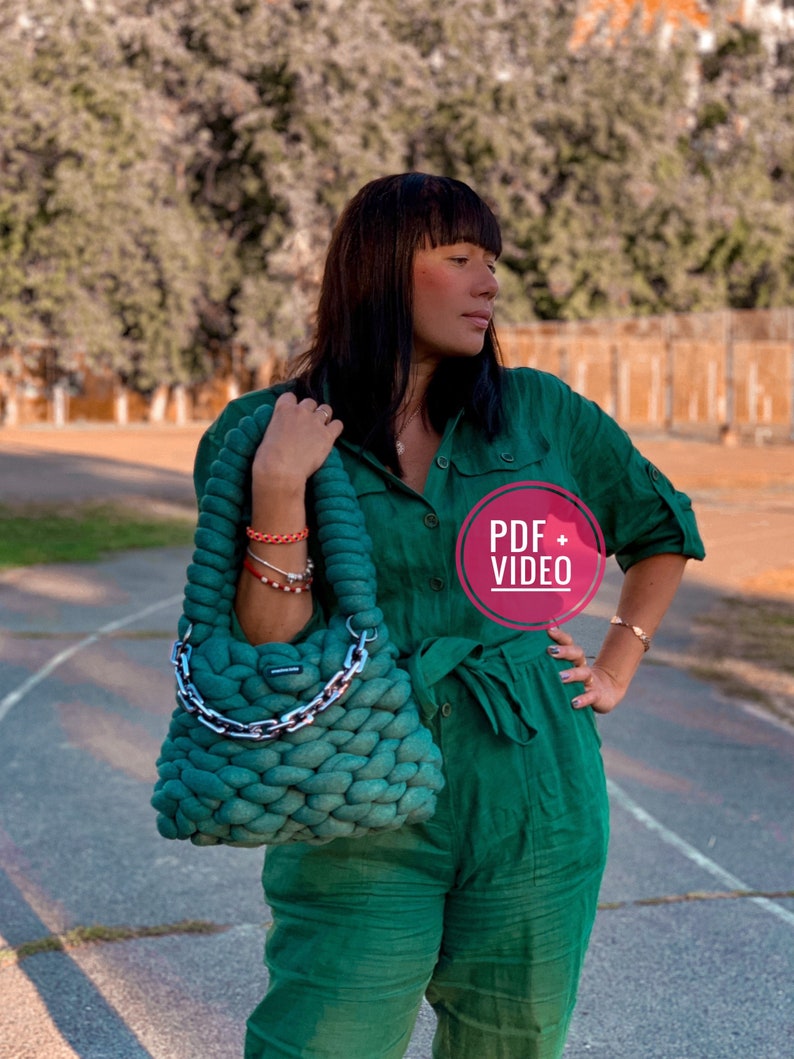 May include: A woman wearing a green jumpsuit holds a green crocheted bag with a silver chain strap. The bag is a chunky knit with a textured surface. The woman is standing on a paved surface with a blurred background of trees.