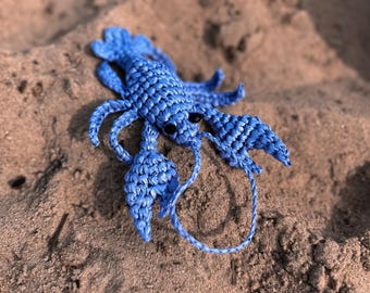 May include: A handmade blue crochet lobster figurine with black eyes, resting on a sandy surface. The lobster has detailed claws, legs, and antennae, showcasing the intricate crochet work. The figurine is a decorative item.