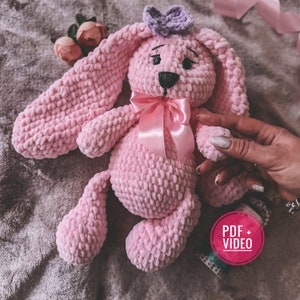 May include: Pink crocheted bunny rabbit toy with a purple bow on its head and a pink satin ribbon around its neck. The bunny is sitting on a gray fuzzy surface. The text "PDF + VIDEO" is visible in the lower right corner of the image.