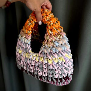 May include: A handmade, colorful tote bag with a triangular shape. The bag is crafted from a knitted fabric in shades of orange, pink, yellow, gray, and purple. Gold metallic thread is woven into the orange handle. The bag is held by a hand.