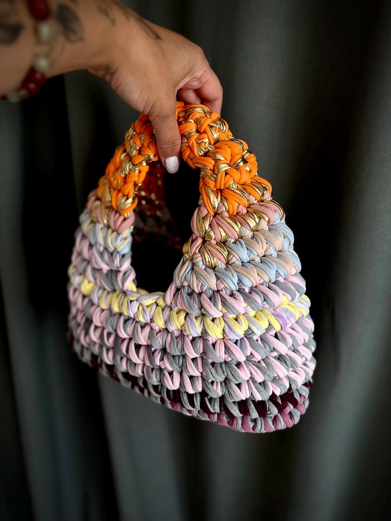 May include: A handmade, colorful tote bag with a triangular shape. The bag is crafted from a knitted fabric in shades of orange, pink, yellow, gray, and purple. Gold metallic thread is woven into the orange handle. The bag is held by a hand.
