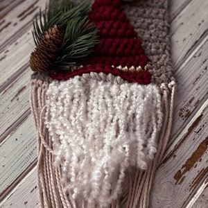 May include: A woven wall hanging featuring a gnome with a red hat, white beard, and a brown pine cone on top. The gnome is made with yarn and has a rustic, handmade look.