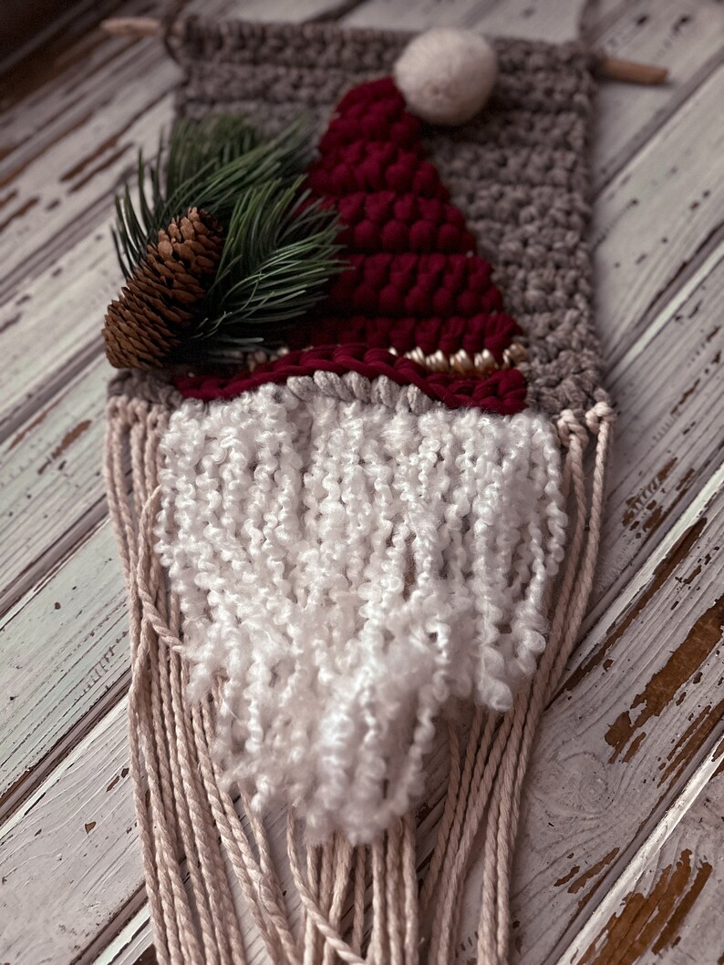 May include: A woven wall hanging featuring a gnome with a red hat, white beard, and a brown pine cone on top. The gnome is made with yarn and has a rustic, handmade look.