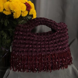 May include: A burgundy crocheted handbag with a braided handle and beaded fringe. The bag is made of a thick, textured yarn with a subtle shimmer. Yellow flowers with red centers are in the background, adding a pop of color.