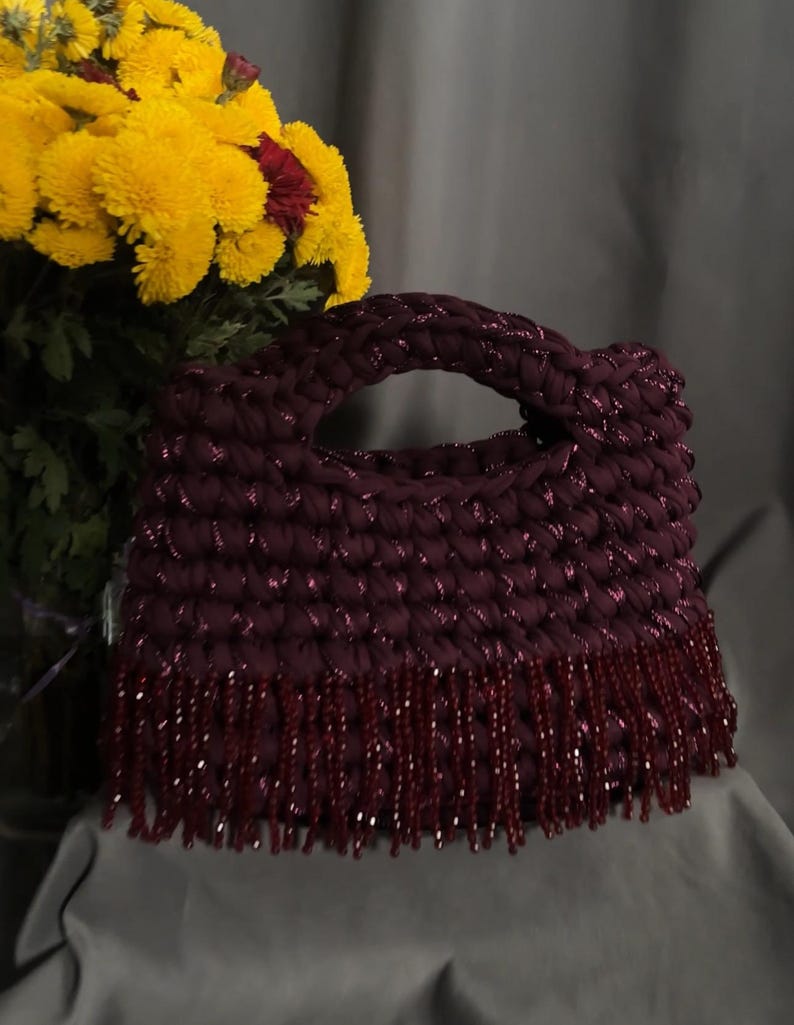 May include: A burgundy crocheted handbag with a braided handle and beaded fringe. The bag is made of a thick, textured yarn with a subtle shimmer. Yellow flowers with red centers are in the background, adding a pop of color.