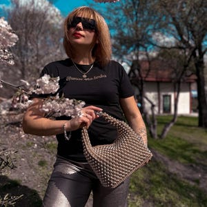 May include: A woman wearing a black t-shirt with the text "@mac.na.torba" in gold lettering, silver sunglasses, a silver necklace, and silver pants. She is holding a beaded handbag.