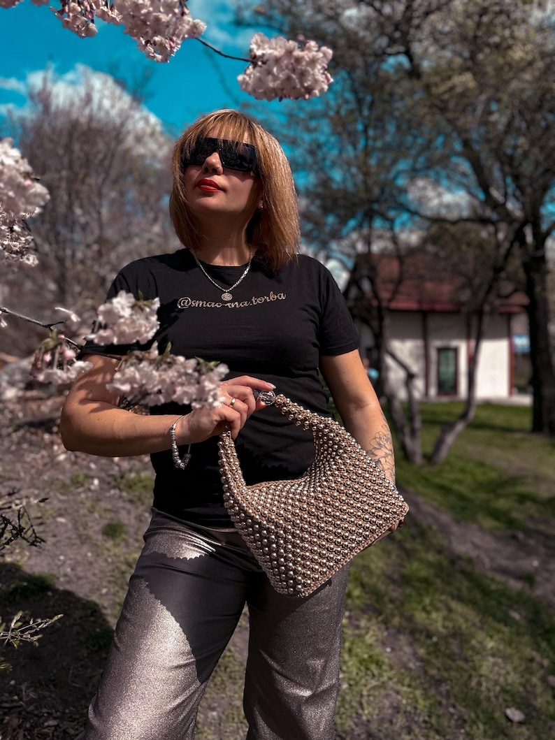 May include: A woman wearing a black t-shirt with the text "@mac.na.torba" in gold lettering, silver sunglasses, a silver necklace, and silver pants. She is holding a beaded handbag.