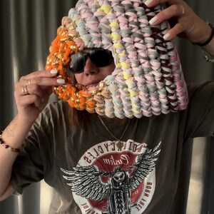 May include: A colorful, handmade head covering made of fabric strips in shades of pink, gray, yellow, and orange. The head covering is worn over the head and face, with sunglasses. The head covering is a unique fashion accessory.