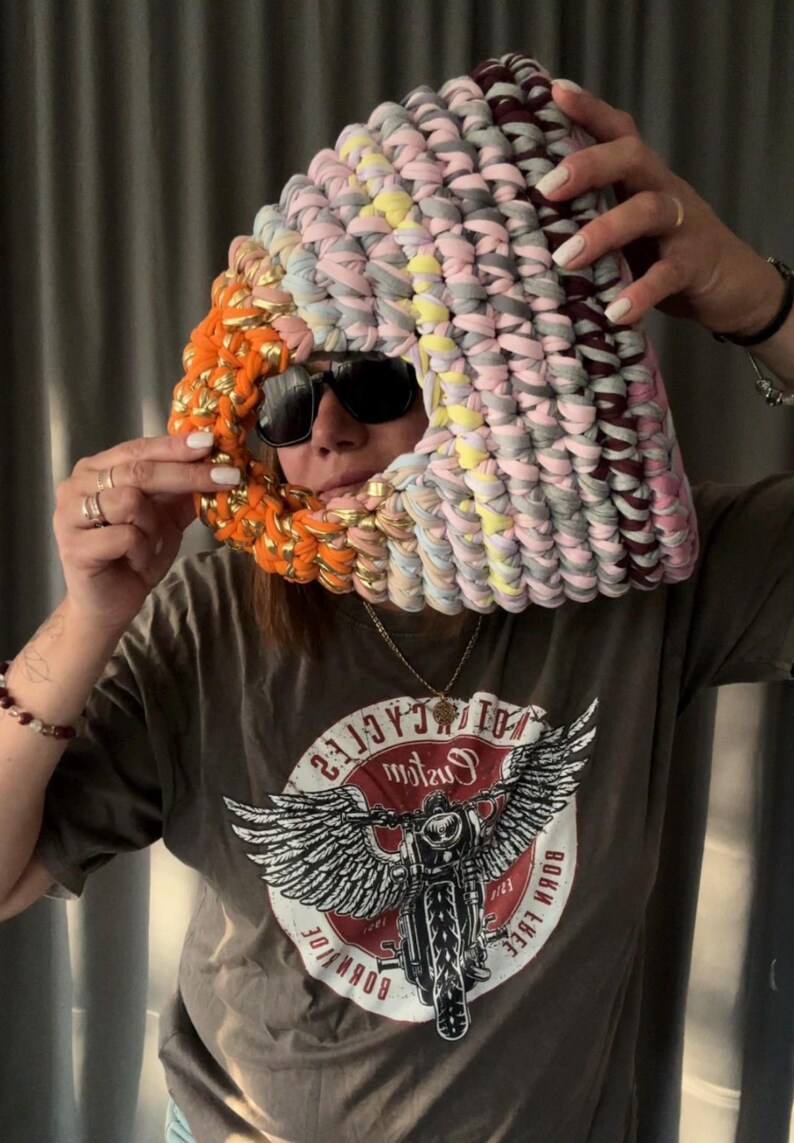 May include: A colorful, handmade head covering made of fabric strips in shades of pink, gray, yellow, and orange. The head covering is worn over the head and face, with sunglasses. The head covering is a unique fashion accessory.