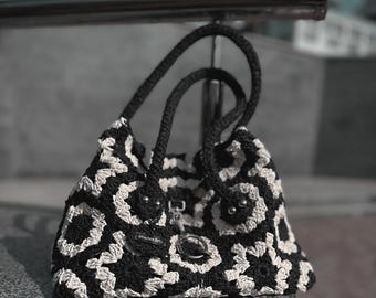May include: A black and white crocheted tote bag with a circular pattern. The bag has a single strap and a metal clasp.
