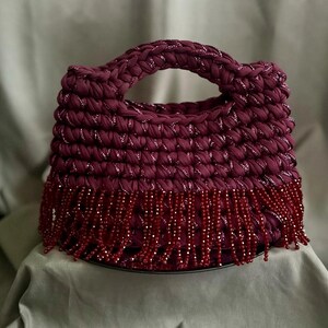 May include: A burgundy crocheted handbag with a rounded handle and beaded fringe. The bag is made of a thick, textured yarn with silver metallic threads woven throughout. The fringe is made of small, dark red beads.